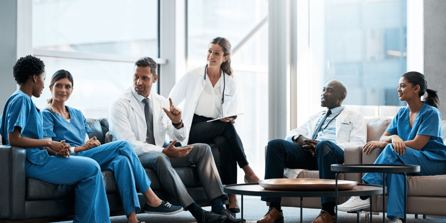 A diverse team of medical doctors and healthcare staff in scrubs sitting together in a bright, modern clinic lounge discussing practice operations and patient scheduling.